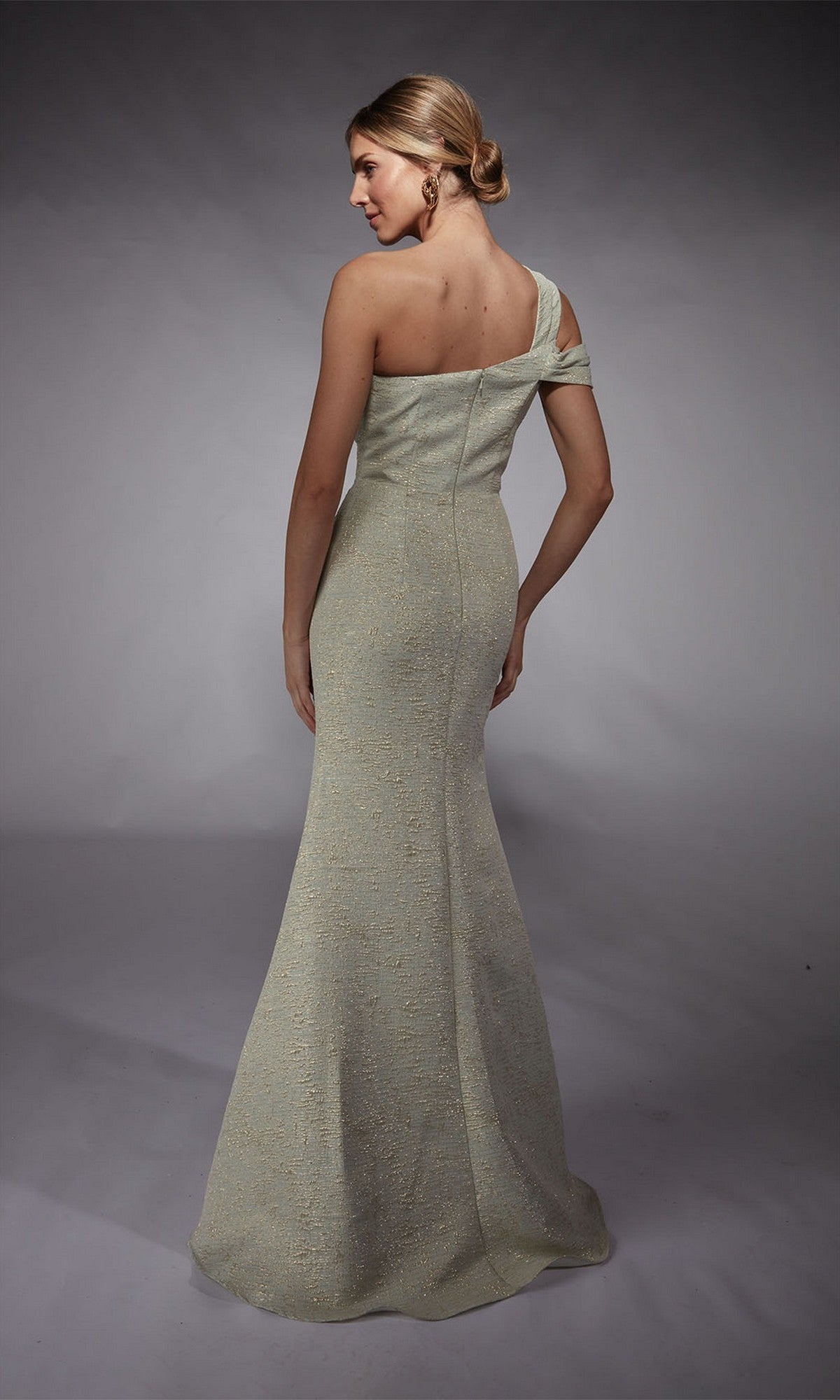 A woman stands with her back to the camera on a plain gray background, wearing the Alyce 27749 long formal dress featuring a light-colored jacquard fabric and a fitted sheath silhouette.